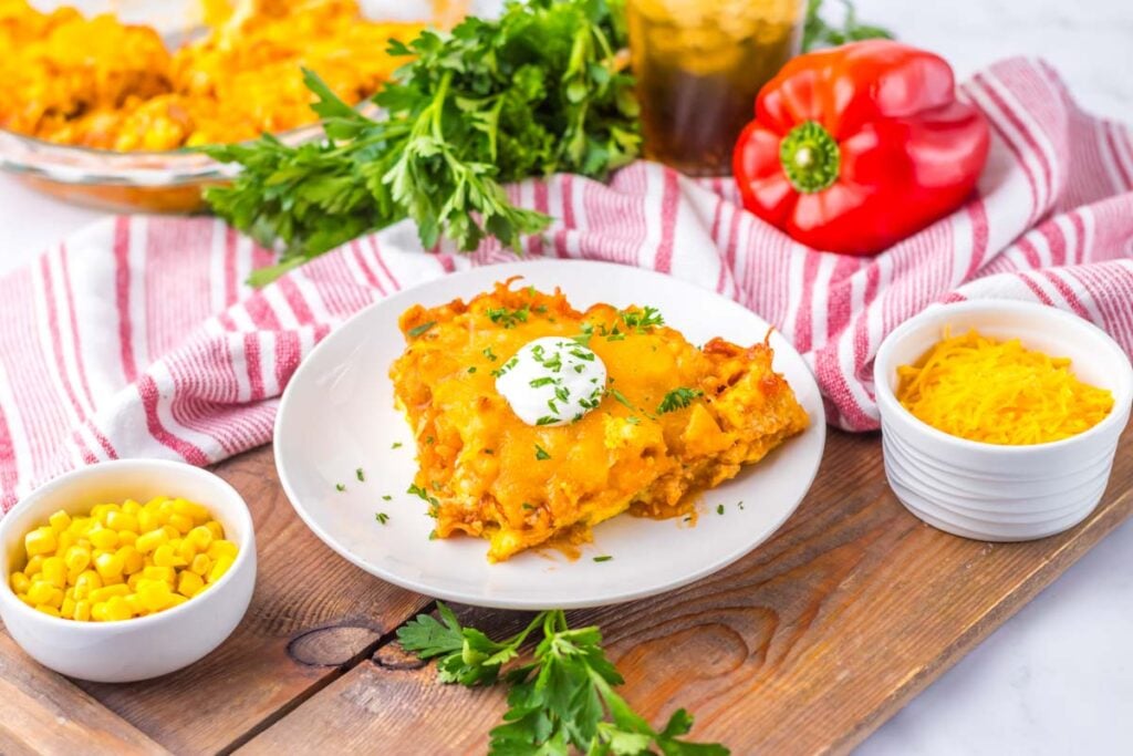 A slice of cheesy chicken tamale pie topped with sour cream and herbs on a white plate, with bowls of corn, shredded cheese, fresh parsley, and a red bell pepper on a wooden board.