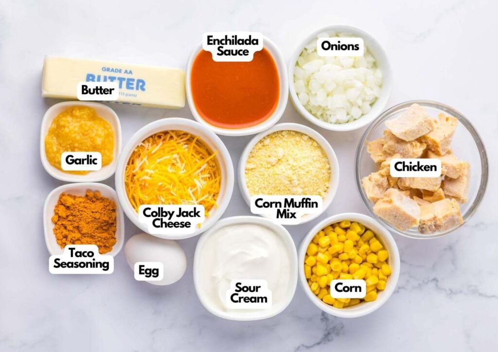 Various labeled ingredients for a chicken tamale pie are arranged in bowls on a marble surface, including chicken, corn, cheese, sour cream, enchilada sauce, onions, and seasonings.