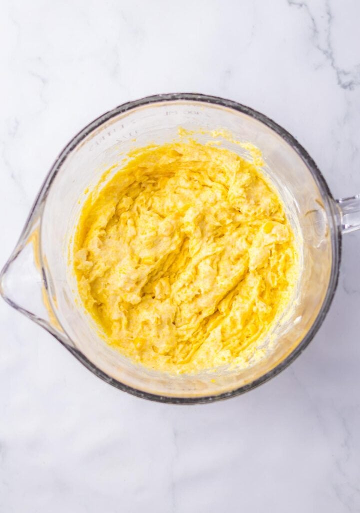 A glass bowl containing a thick, creamy yellow chicken tamale pie batter with visible streaks and texture, viewed from above on a marble surface.