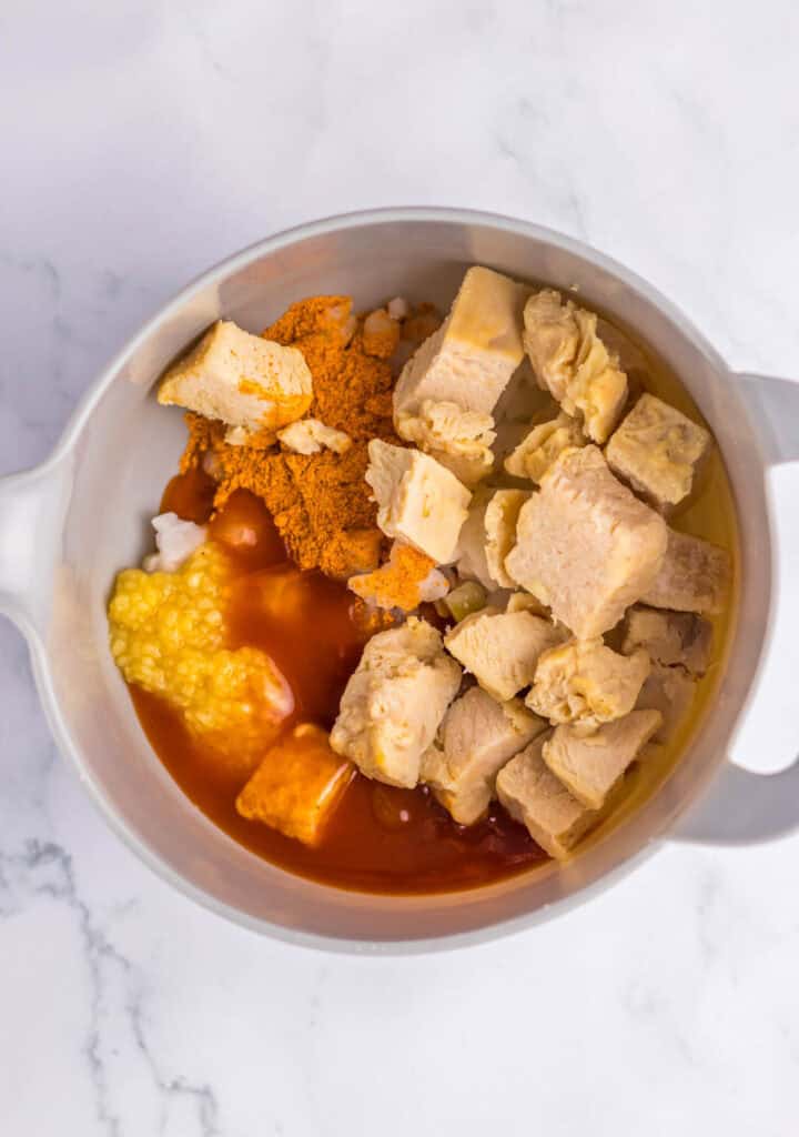 A bowl of ingredients for a chicken tamale pie with chunks of chicken, vegetables, and a rich, spicy sauce, viewed from above on a marble surface.