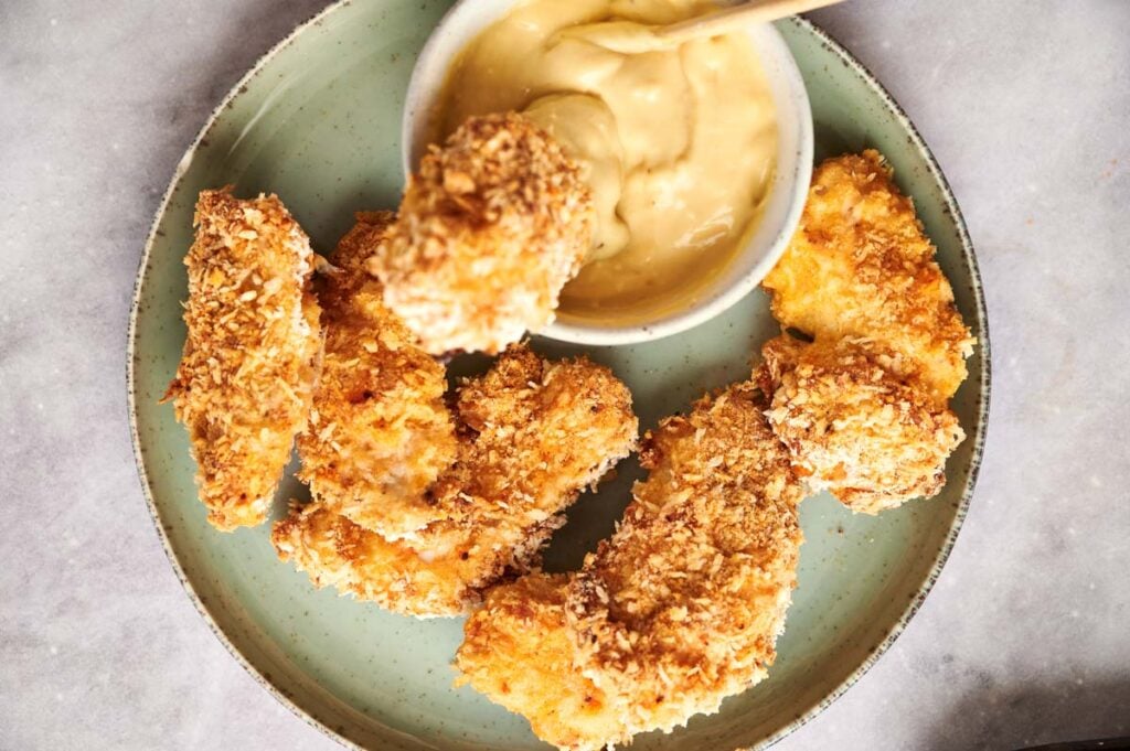 A plate of crispy chicken tenders with a small bowl of creamy dipping sauce, one tender being dipped in for the perfect bite.
