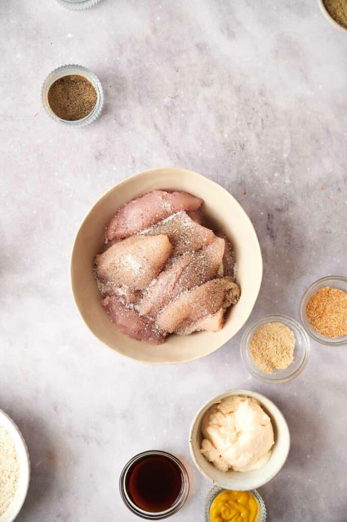 Raw chicken tenders being seasoned, surrounded by various ingredients and spices.
