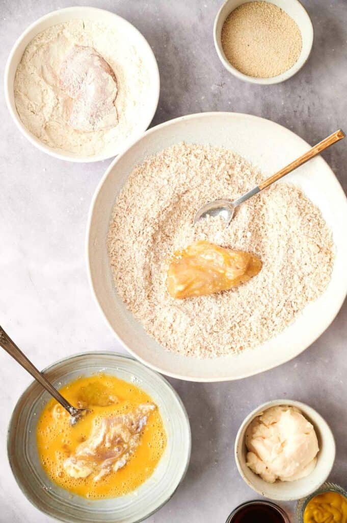 Ingredients for baking arranged on a table with flour, eggs, chicken tenders, and other items in separate bowls.