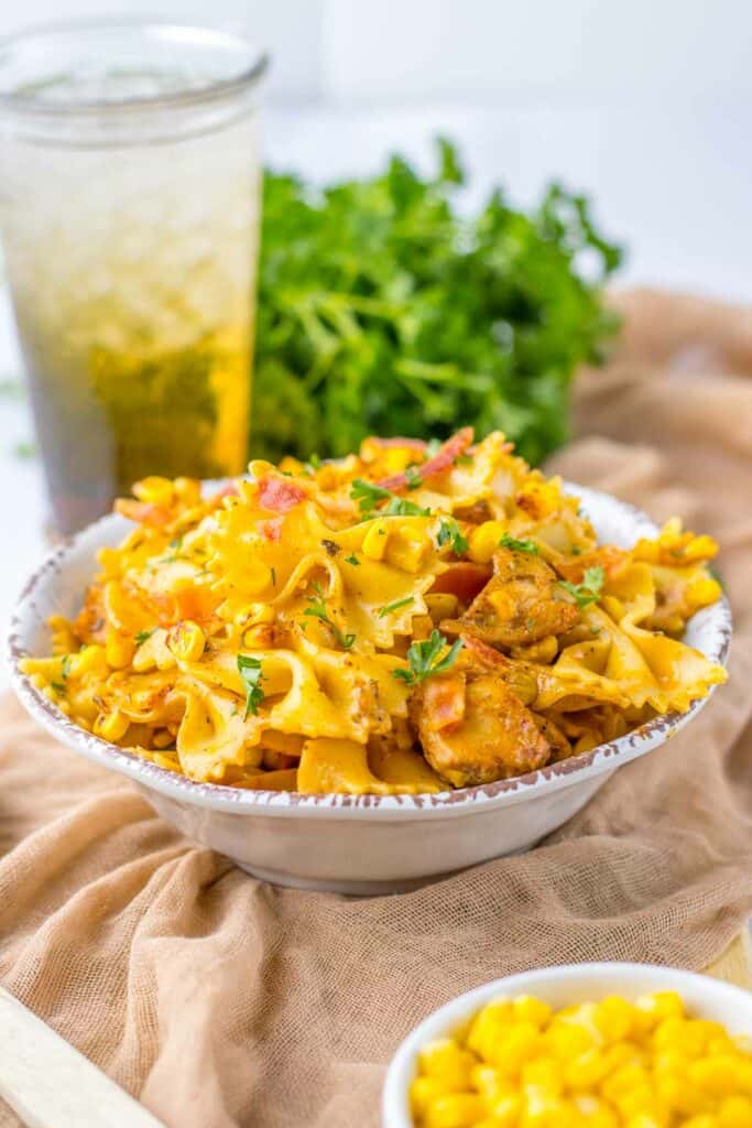 A bowl of chicken and corn pasta garnished with herbs, served with a side of corn and a glass of iced tea.