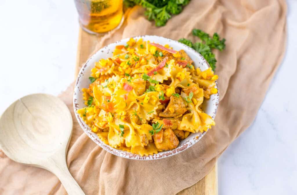 Bowl of creamy chicken and corn pasta topped with herbs on a marble surface, accompanied by a wooden spoon and a glass of iced tea.