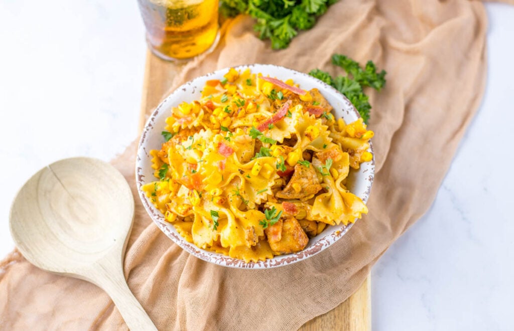 A bowl of Chicken and Corn Pasta with chunks of meat, diced vegetables, and herbs, placed on a beige cloth next to a wooden spoon and a glass of drink.