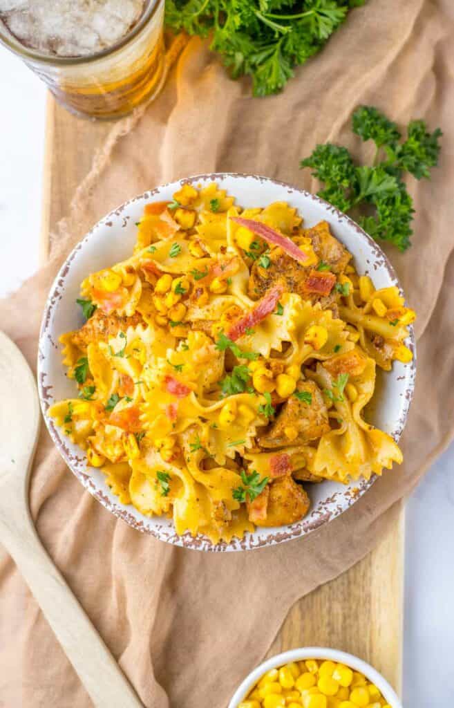 A bowl of chicken and corn pasta topped with bacon bits and chicken, served on a rustic tablecloth, with a glass of beer and fresh parsley nearby.
