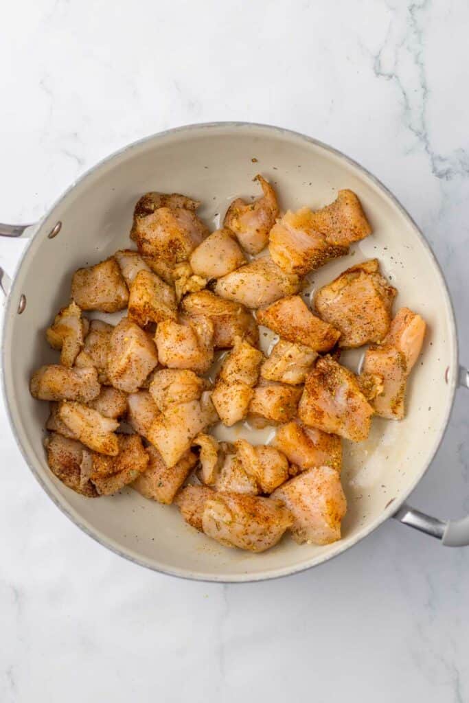 Raw chicken and corn pasta seasoned with spices in a white cooking pot on a marble surface.