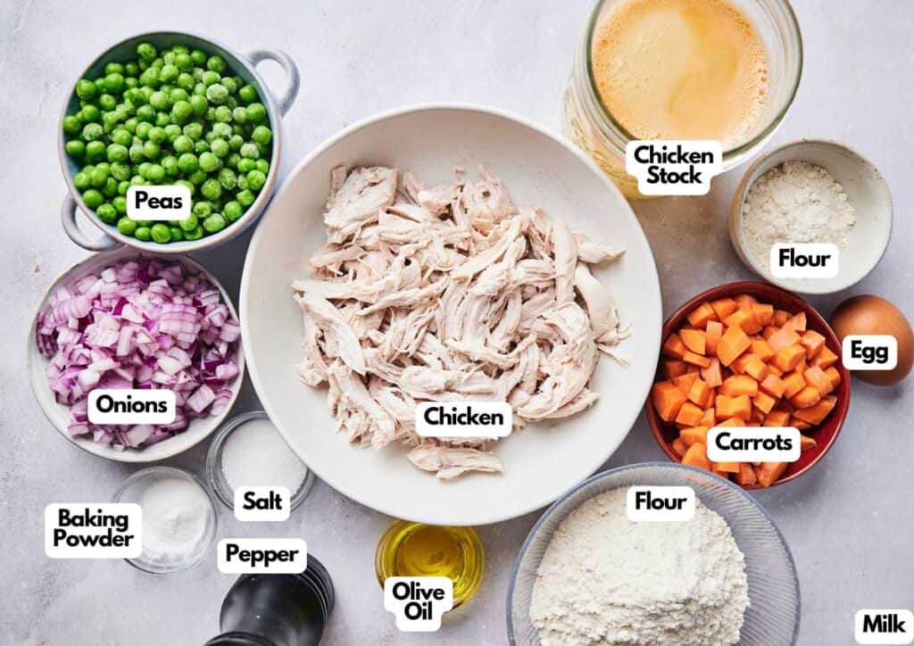 Ingredients for a recipe arranged on a table, including shredded chicken, peas, onions, carrots, flour, egg, milk, chicken stock, olive oil, baking powder, salt, and pepper—perfect essentials for a hearty chicken and dumpling casserole.