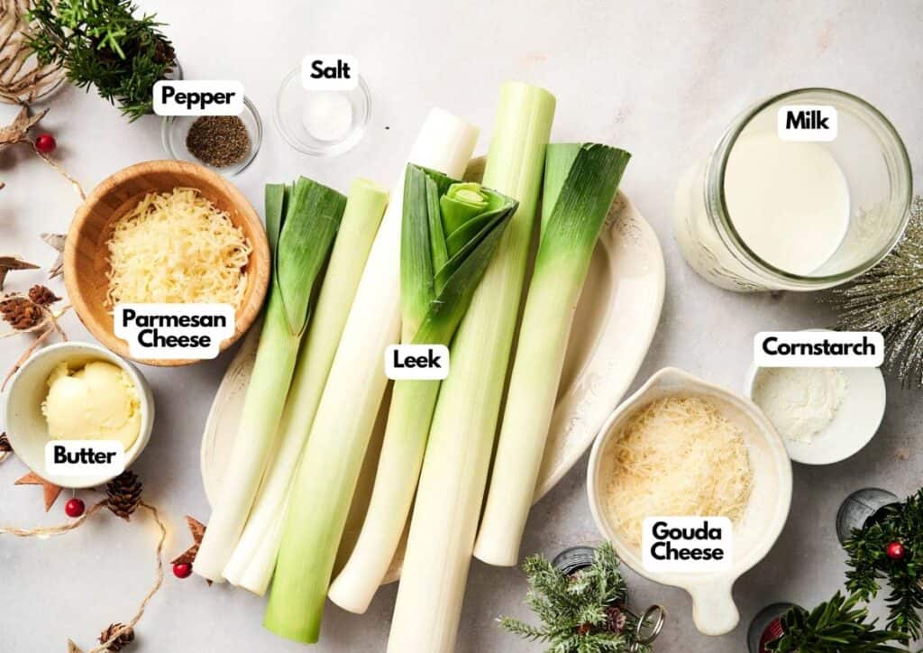 Top-down view of ingredients for a Christmas Casserole leek dish, including leeks, parmesan cheese, gouda cheese, butter, salt, pepper, milk, and cornstarch, all labeled on a white surface.