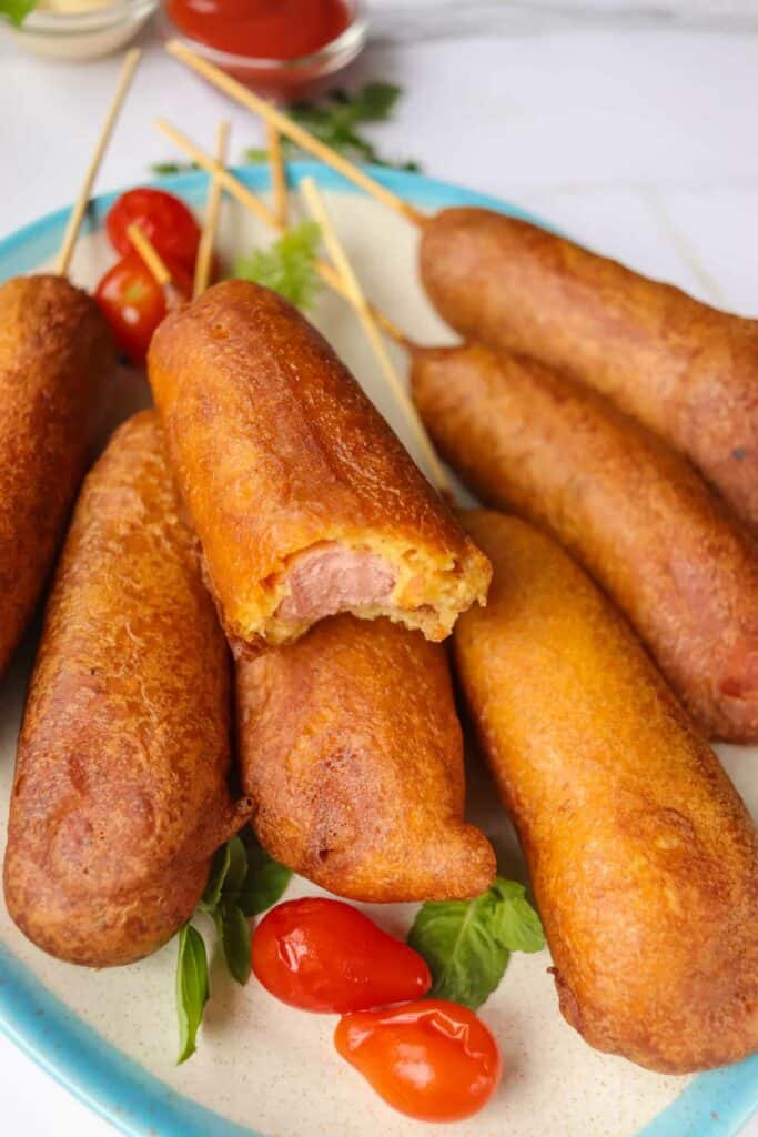 Plate of corn dogs, one bitten, with cherry tomatoes and garnishes on a white background.