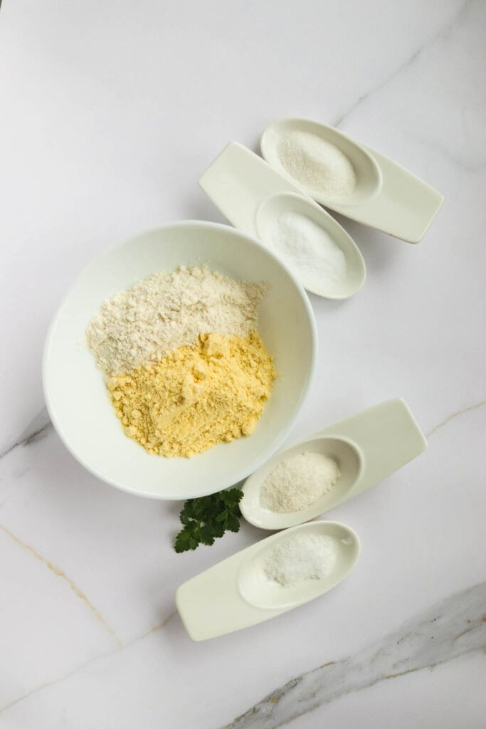 Three types of flour in white bowls on a marble surface, with small white scoops and a sprig of parsley beside corn dogs.