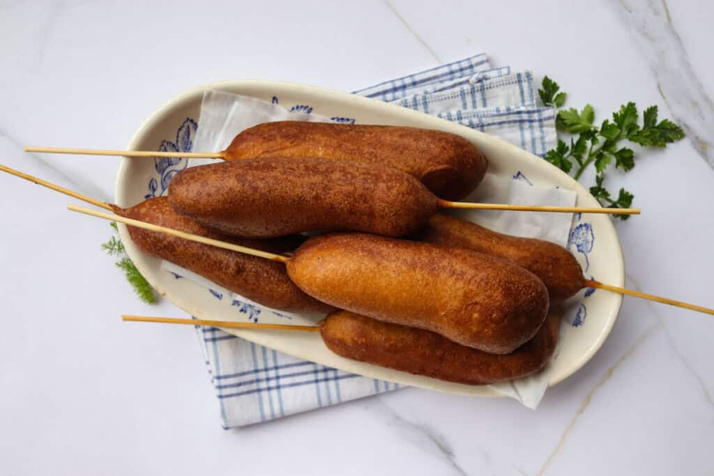 Four corn dogs on a skewer, resting on an oval plate with a blue patterned cloth, on a marble surface.