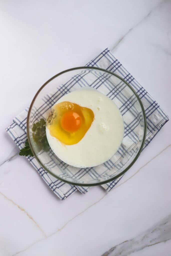 An egg in a clear glass dish placed next to corn dogs on a blue and white checkered cloth on a marble surface.