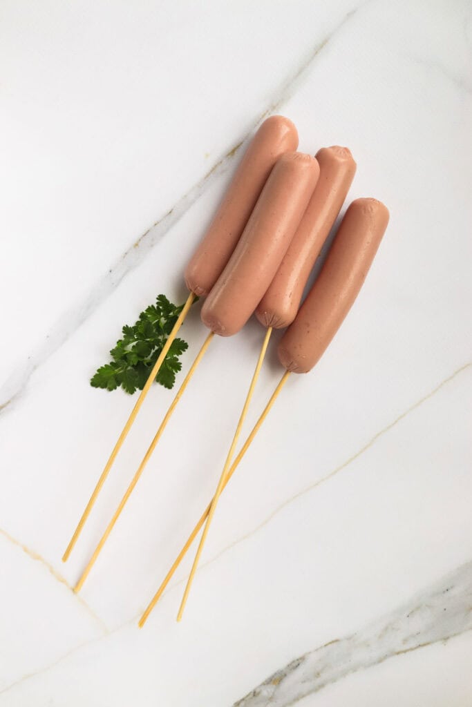 Four corn dogs skewered on sticks with a sprig of parsley, arranged on a marble surface.