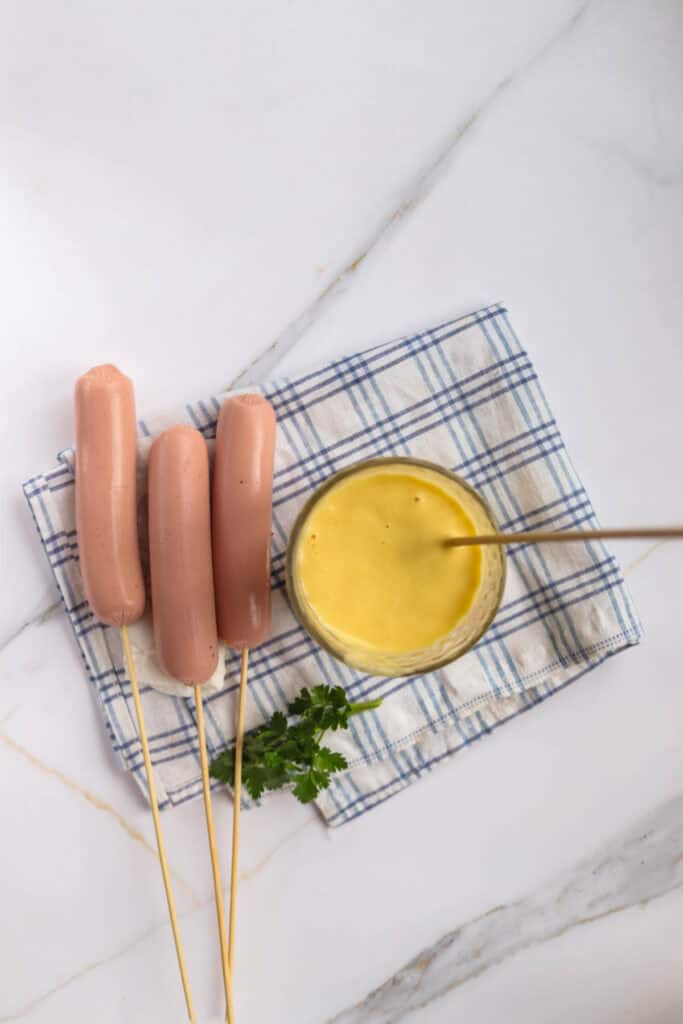 Three corn dogs on wooden skewers next to a bowl of mustard and a sprig of parsley on a blue and white checked napkin, all placed on a marble surface.