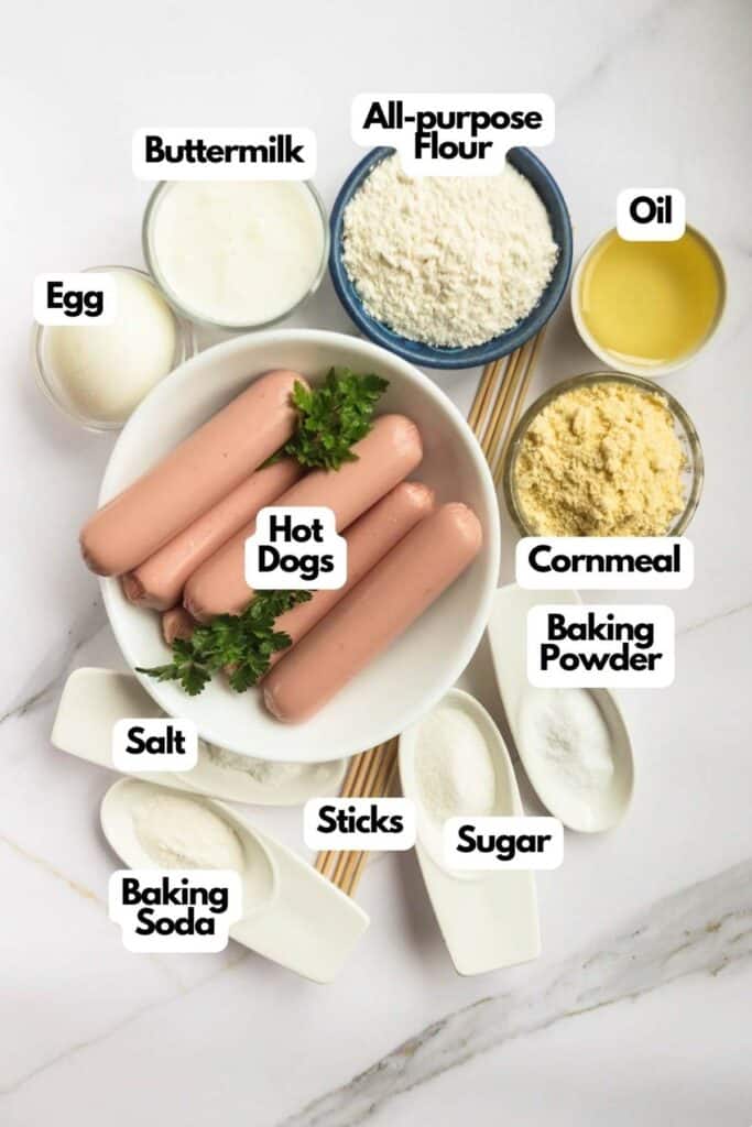 Ingredients for a recipe laid out on a marble surface, including corn dogs, egg, buttermilk, various baking powders and sugars, each labeled.