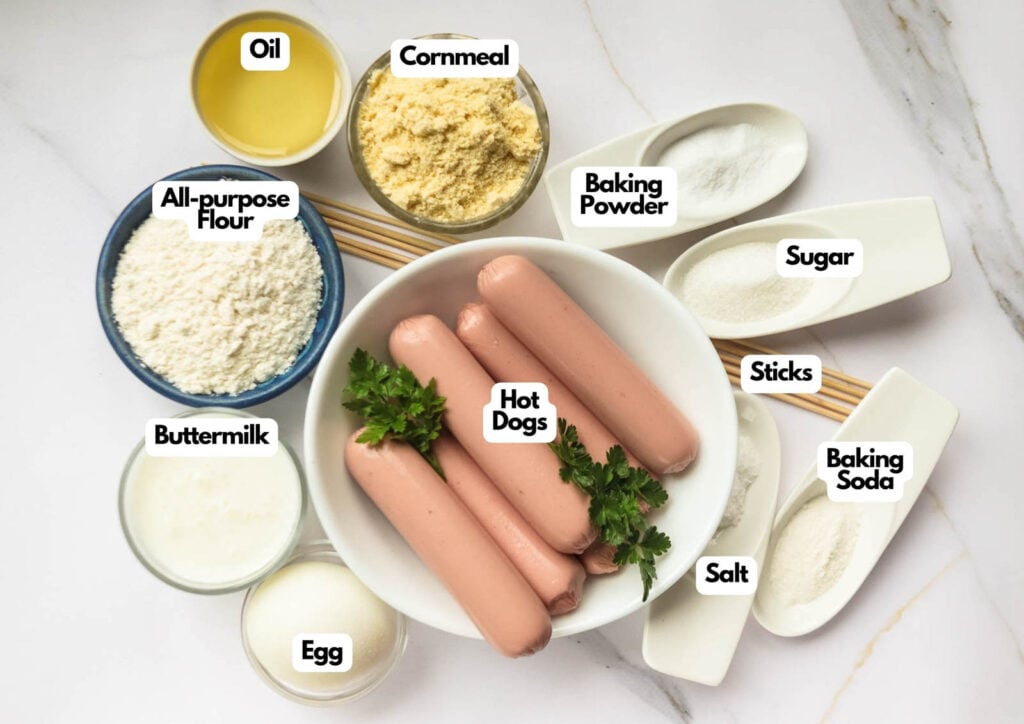 Assorted corn dogs ingredients, including hot dogs, cornmeal, flour, oil, buttermilk, egg, baking powder, baking soda, sugar, salt, and wooden sticks, neatly arranged on a counter for making classic corn dogs.