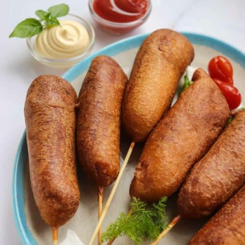 A Plate of corn dogs with dipping sauces: ketchup and mayonnaise, garnished with fresh herbs and cherry tomatoes on a marble surface.