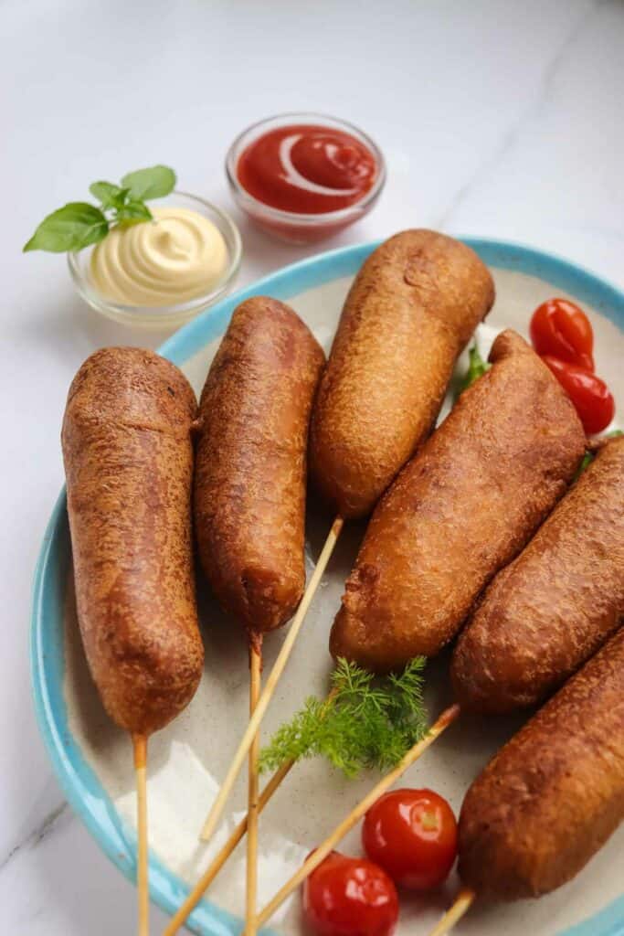 A Plate of corn dogs with dipping sauces: ketchup and mayonnaise, garnished with fresh herbs and cherry tomatoes on a marble surface.