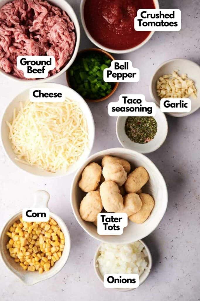 Ingredients for a cowboy casserole recipe laid out on a table, including ground beef, crushed tomatoes, cheese, bell pepper, taco seasoning, garlic, corn, tater tots, and onions