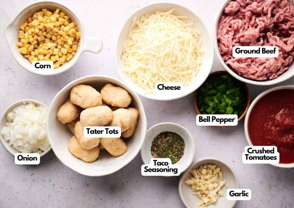 Bowls of ingredients for cowboy casserole, including corn, shredded cheese, ground beef, diced bell pepper, tater tots, chopped onion, taco seasoning, garlic, and crushed tomatoes on a white surface.