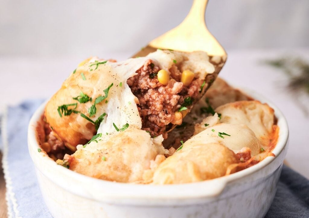 A spoon serving a portion of cowboy casserole with ground meat, corn, melted cheese, and chopped herbs in a white dish.