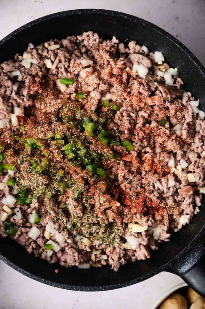 Ground meat cooking in a skillet with diced onions and green peppers, seasoned with red and green spices for a cowboy casserole.