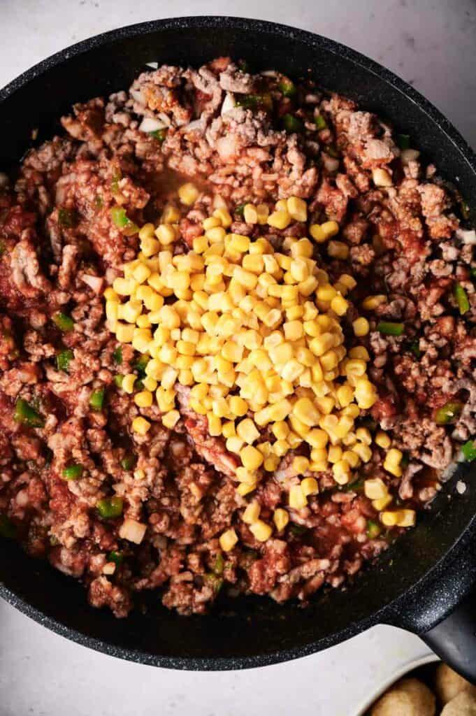 Ground meat and green peppers cooking in a skillet with a pile of corn added in the center, creating a cowboy casserole.