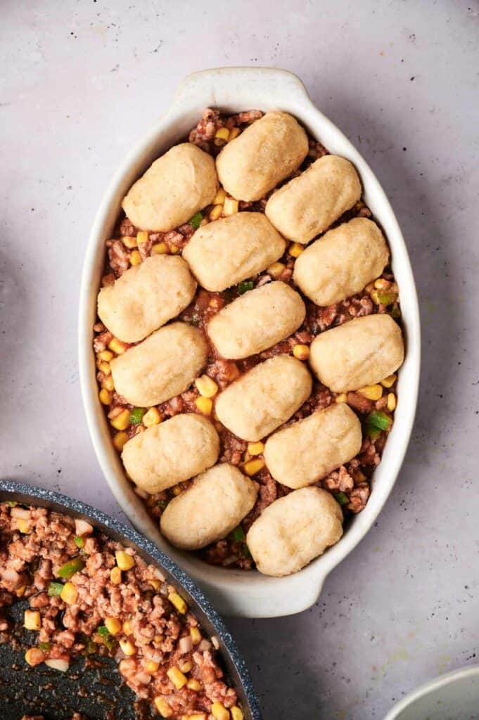 A cowboy casserole dish containing a beef and vegetable mixture topped with biscuit dough, photographed from above on a light surface.