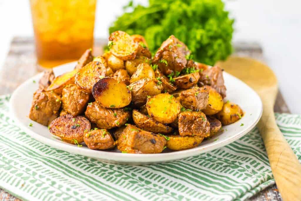 A plate of honey chipotle steak and potatoes garnished with herbs on a green and white striped cloth, with a glass of iced tea in the background.
