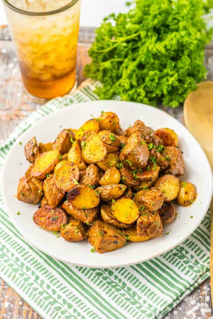 A plate of honey chipotle steak and potatoes garnished with herbs, accompanied by a glass of iced tea and fresh parsley on the side, placed on a green striped napkin.