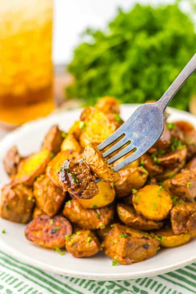 A fork piercing a seasoned honey chipotle steak and potatoes on a plate full of honey chipotle roasted potatoes, with fresh herbs and a drink in the background.