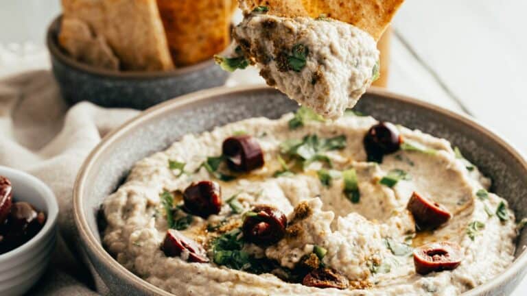 A bowl of hummus topped with olives and herbs, with a piece of pita bread being dipped into it.
