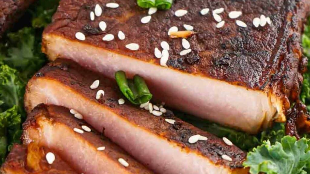 Close-up of sliced asian style pork chops garnished with sesame seeds and green chili, served with a side of kale.