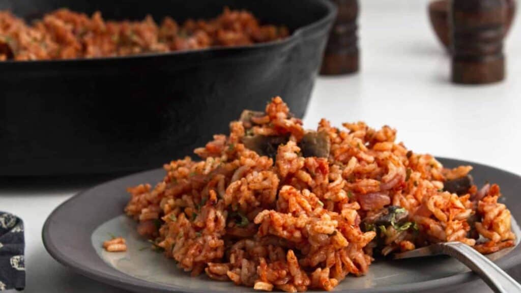 A plate of jollof rice with a spoon, featuring mixed vegetables, served beside a cast iron skillet on a white table.