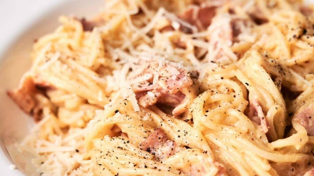 A close-up image of a creamy spaghetti carbonara garnished with grated cheese and black pepper, served on a white plate.