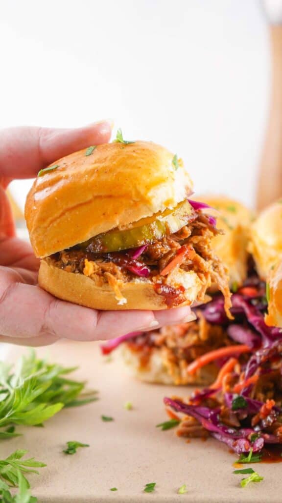 A hand holds pulled pork sliders with pickles and slaw.