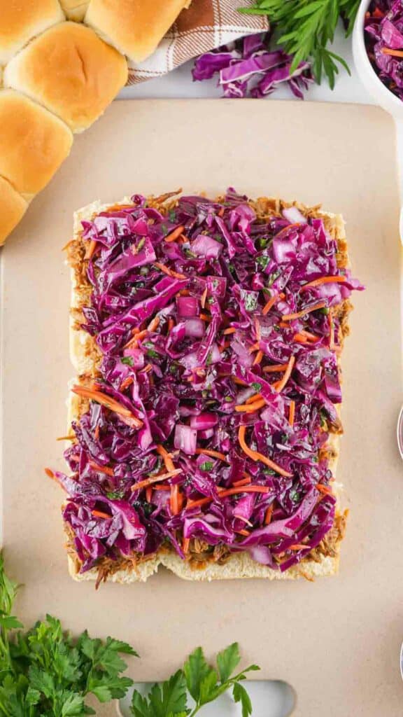 A serving of red cabbage slaw on a rectangular dish, accompanied by pulled pork sliders, surrounded by fresh ingredients and dinner rolls.