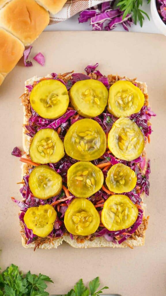 A colorful vegetable sandwich with pickles on top and pulled pork sliders on a light background.