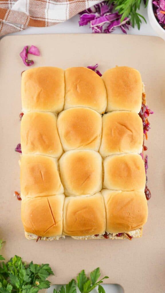 A square sandwich made with a large block of linked dinner rolls, filled with a layer of salad and pulled pork, and served on a beige surface.