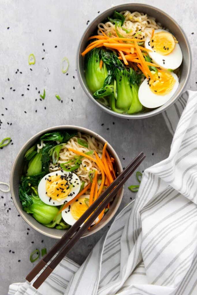 Two bowls of elevated ramen noodles with boiled eggs, vegetables, and a pair of chopsticks on a gray surface.