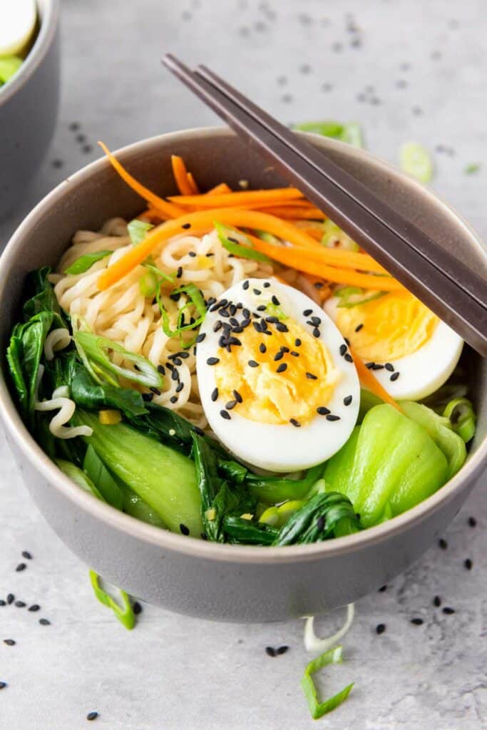 An elevated bowl of ramen noodle soup with spinach, carrots, a boiled egg, and sprinkled with black sesame seeds, accompanied by chopsticks.