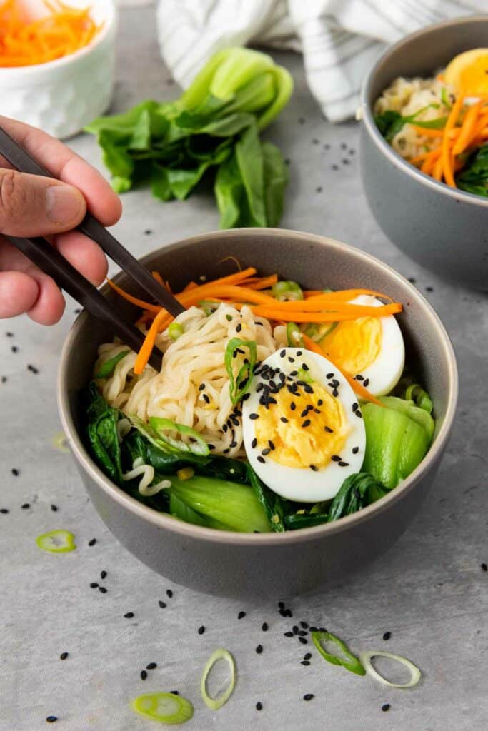 Hand holding chopsticks over a bowl of elevated ramen noodles with soft-boiled egg, vegetables, and sesame seeds.