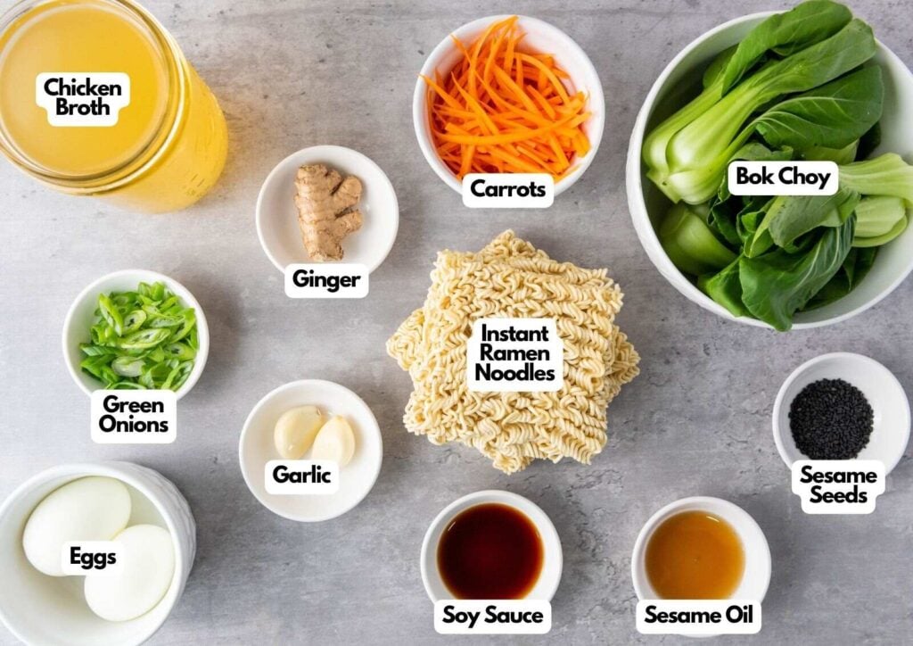 Ingredients for elevated ramen noodles laid out on a table: chicken broth, carrots, bok choy, green onions, ginger, garlic, eggs, instant ramen noodles, soy sauce, sesame oil, and sesame seeds.