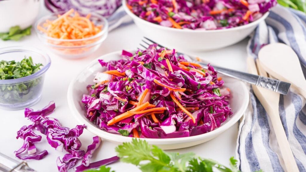 A bowl of Red Cabbage Coleslaw made with shredded cabbage, carrots, and herbs sits on a table with utensils, a striped napkin, and small bowls of chopped vegetables.