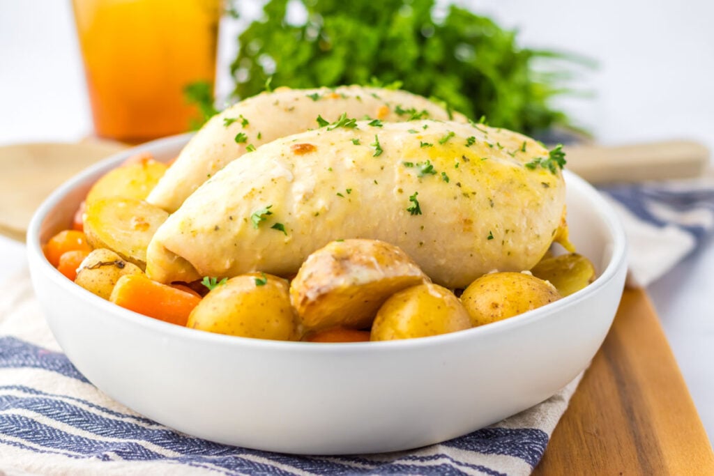 A bowl of slow cooker ranch chicken breasts garnished with herbs, served alongside roasted potatoes and carrots on a table with a striped napkin.