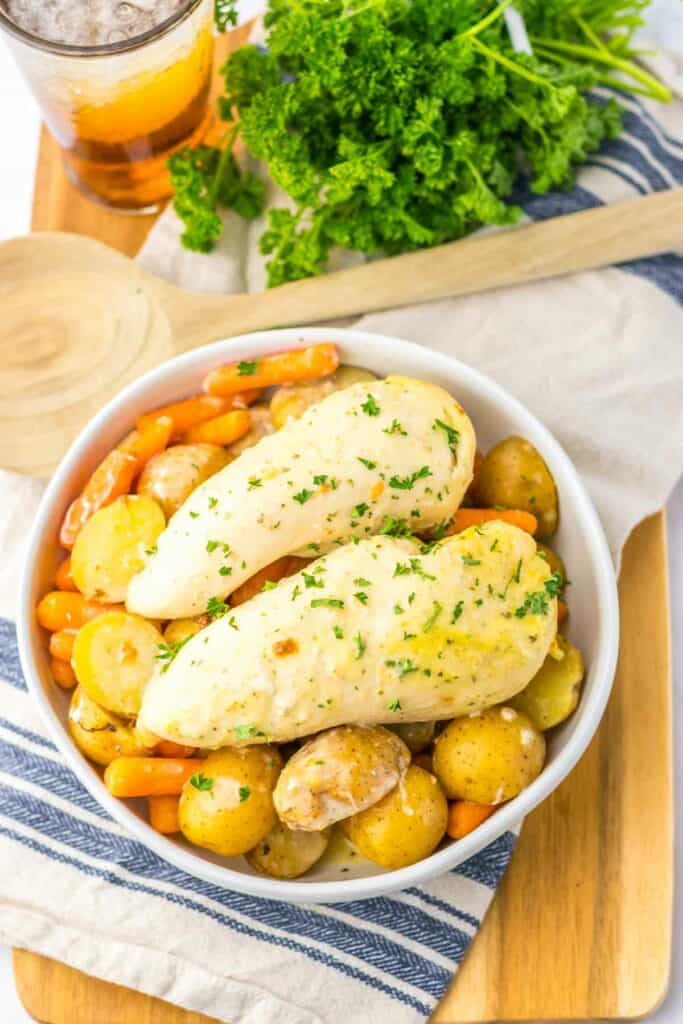 A dish of slow cooker ranch chicken breasts served with carrots and potatoes, garnished with parsley, presented on a wooden tray with a glass of iced tea.