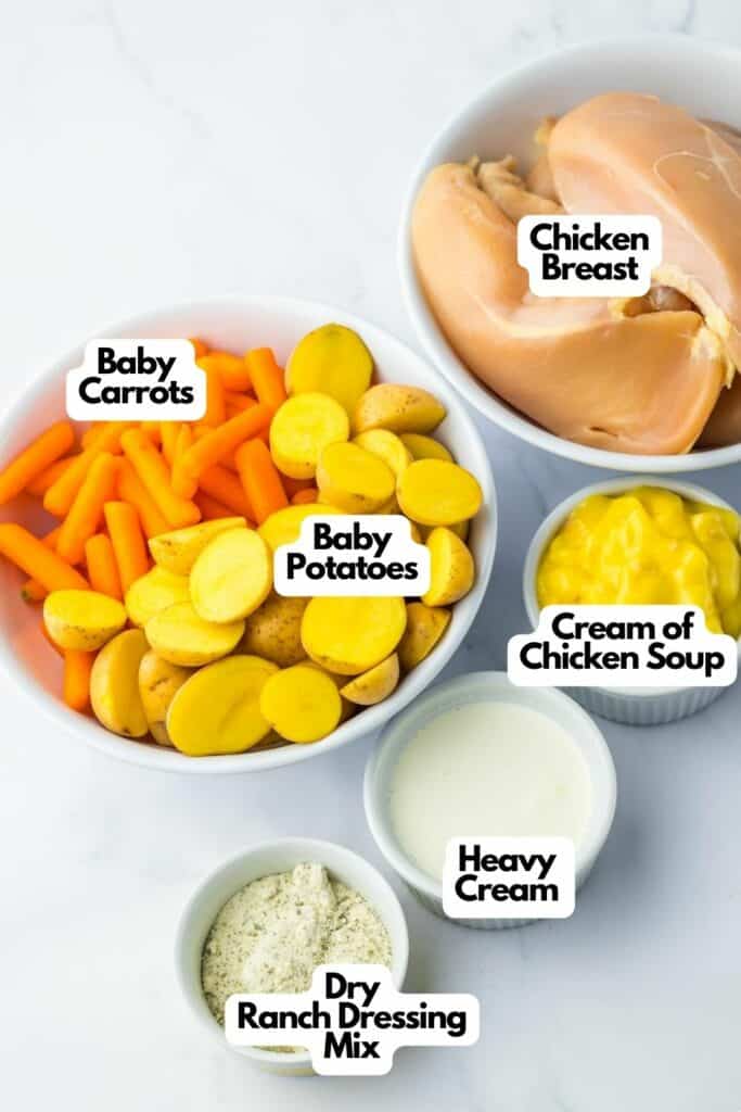 Ingredients for a slow cooker ranch chicken recipe labeled on a white countertop, including chicken breast, baby carrots, baby potatoes, cream of chicken soup, heavy cream, and dry ranch dressing mix.