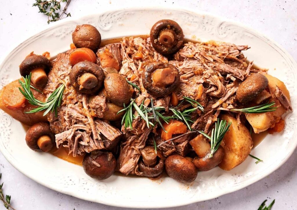 Shredded slow cooker pot roast served on a white platter with whole mushrooms, potatoes, carrots, and sprigs of fresh rosemary.